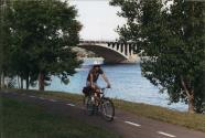 Randonnée cycliste. Un participant sur son vélo sur la piste cyclable sur le bord de la rivière ; on voit un pont également. [1987].