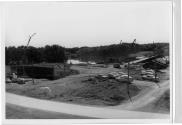 Construction de l'autoroute 15 (Autoroute des Laurentides) sur le bord d'un cours d'eau avec machinerie lourde, voitures et bâtiments