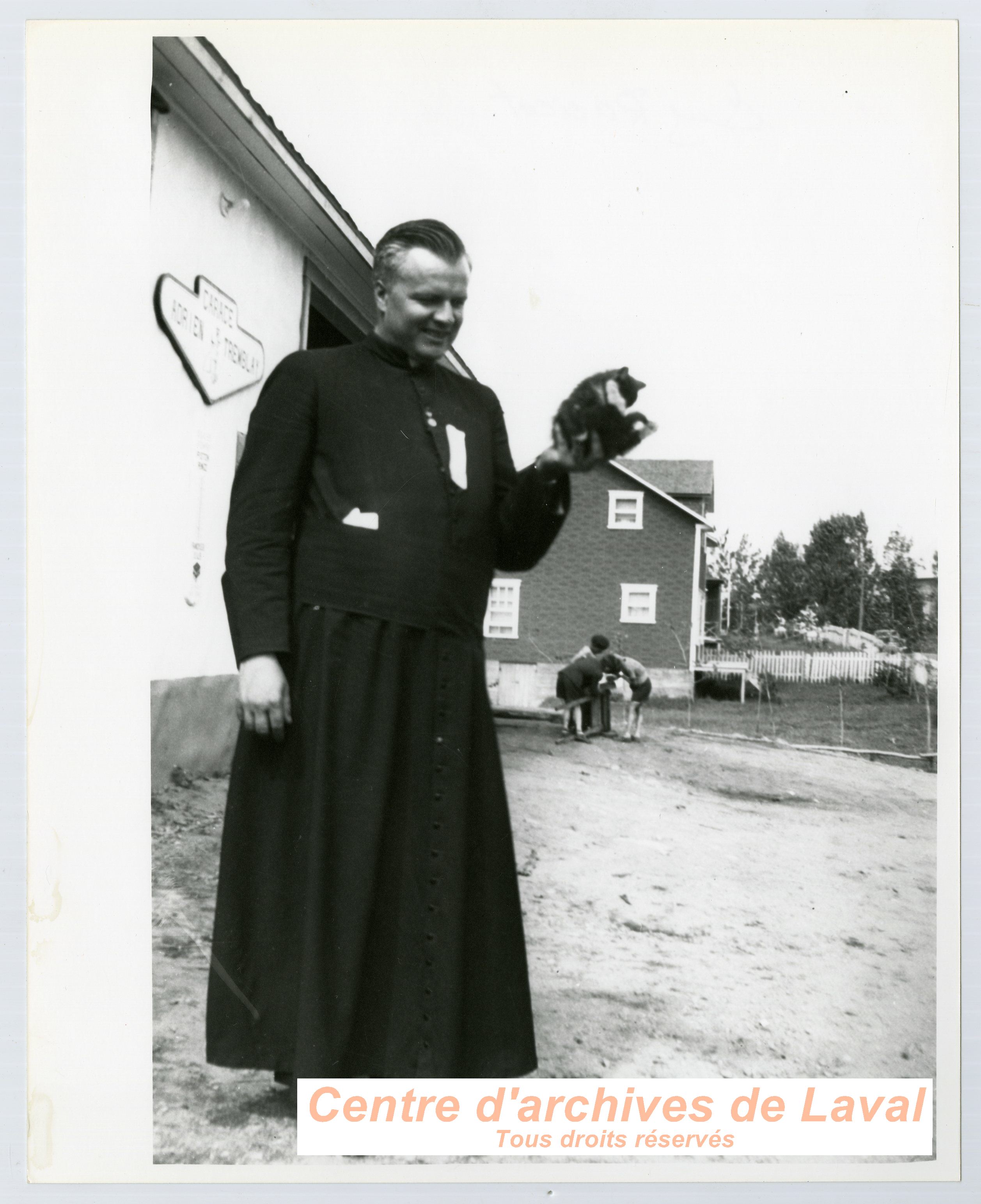 Le chanoine Guy Racicot tenant un chaton devant le garage Adrien Tremblay  Saint-Benot.